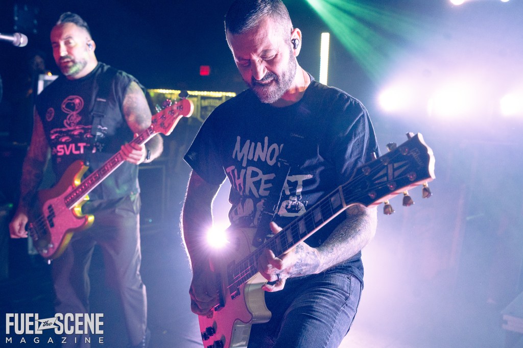 A close-up image of a band performance, featuring a guitarist intensely playing his instrument under colorful stage lights, while a bassist is partially visible in the background.