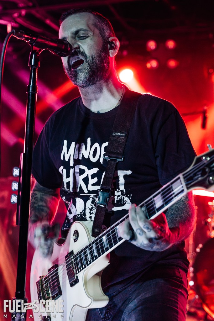 A musician passionately performing on stage with a microphone and electric guitar, wearing a black shirt with a graphic design, illuminated by vibrant red stage lights.