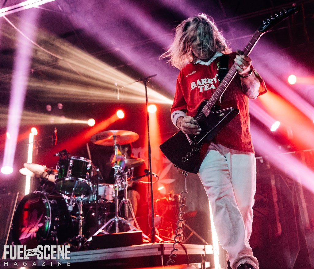 A guitarist performing energetically on stage with vibrant lights in the background during a live concert.