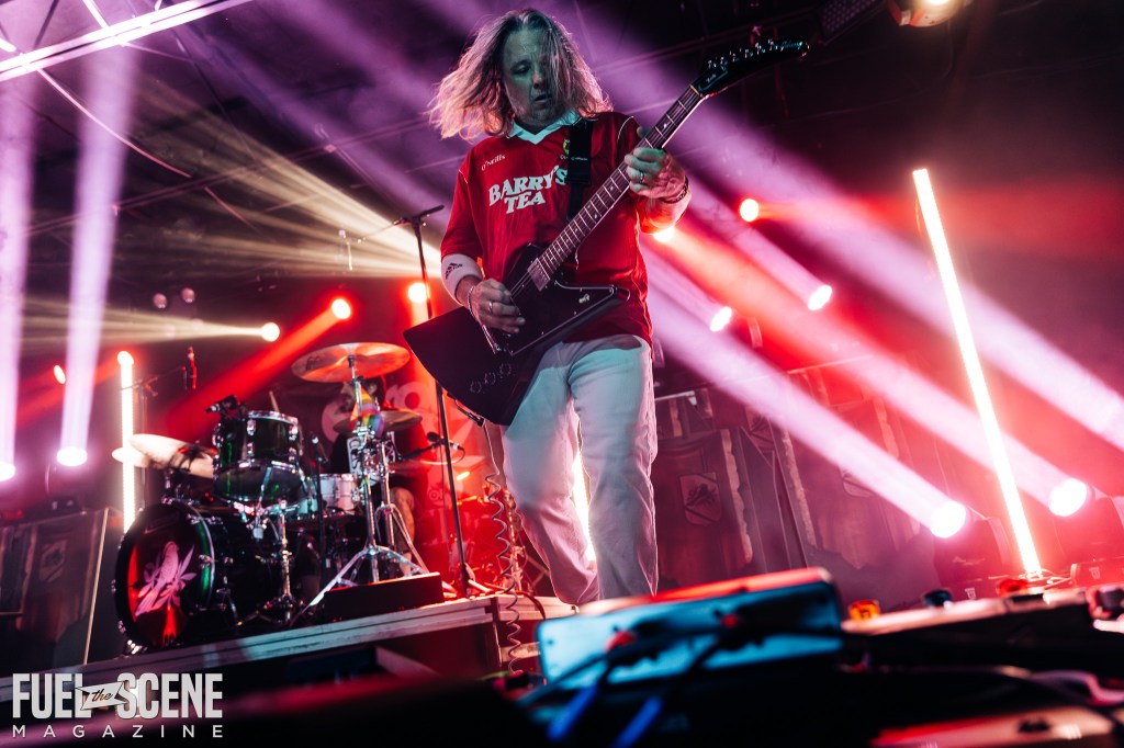 A guitar player on stage with long hair and a red shirt, performing passionately in front of a lit-up backdrop during a concert.