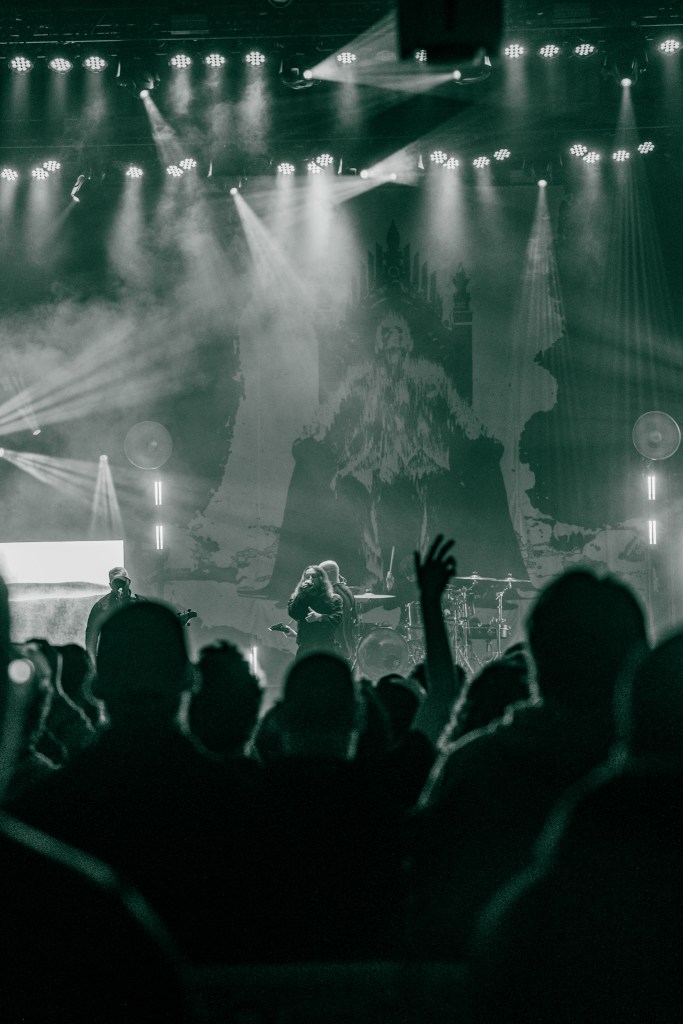 A crowd of fans at a metal concert with their hands raised, illuminated by stage lights, while a band performs in the background. A large graphic banner is displayed behind the musicians.