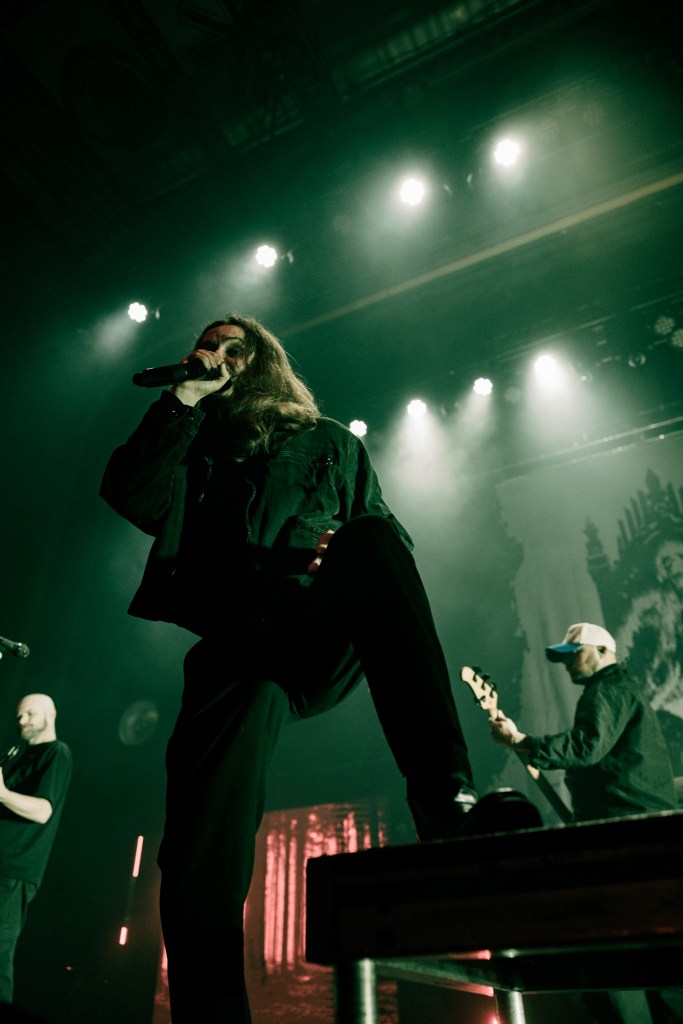 A vocalist with long hair passionately singing into a microphone on stage, illuminated by stage lights, with a guitarist in the background.