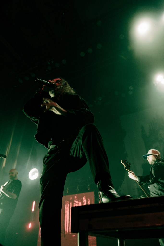 A musician passionately performing on stage, holding a microphone with a dramatic pose, surrounded by green lighting.