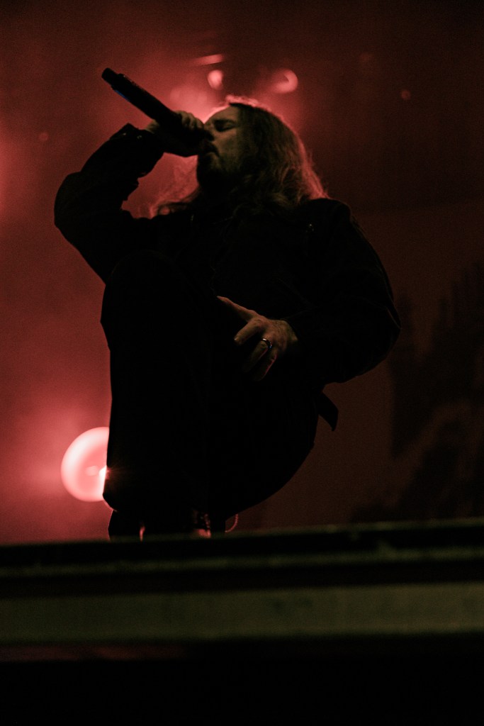 A performer on stage, passionately singing into a microphone, with dramatic red lighting illuminating the scene.