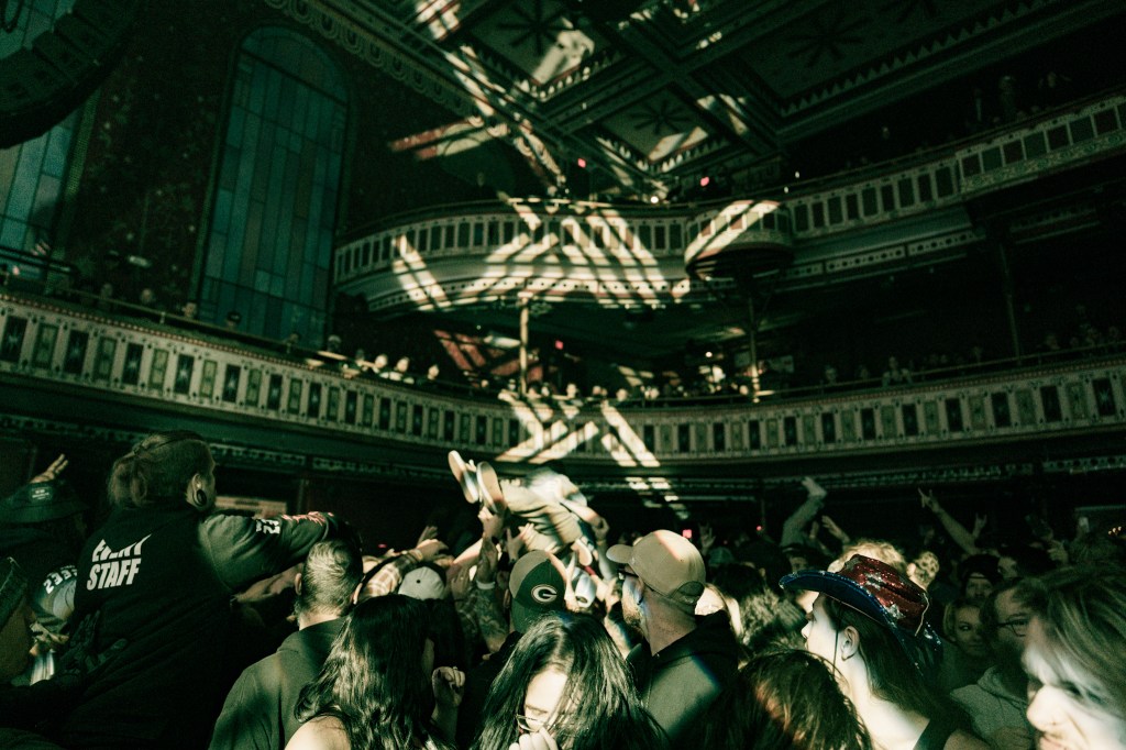 Crowd at a live music event with people crowd-surfing and energetic engagement, illuminated by stage lights in The Tabernacle.