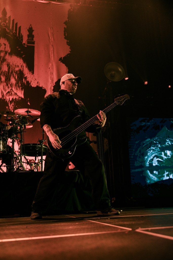 A bassist performing energetically on stage during a concert, wearing a cap and black attire, with dramatic stage lighting and a backdrop featuring bold graphics.