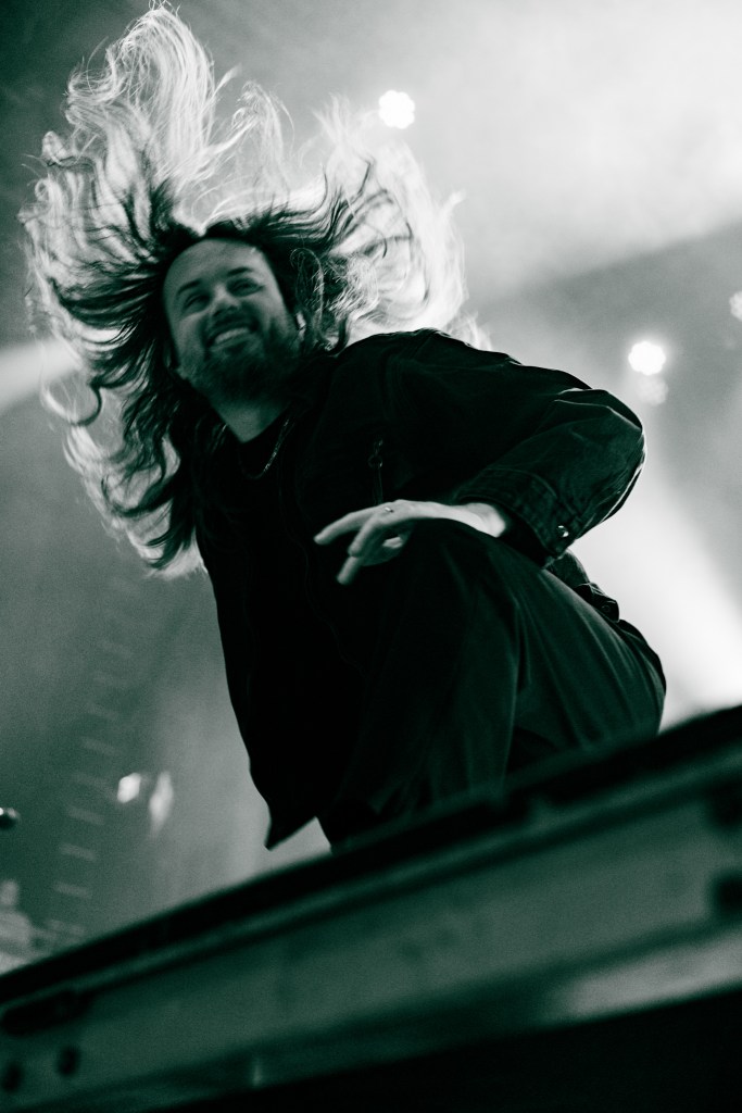 A musician in a black outfit performs energetically on stage, with long hair flowing as he moves, captured in a dramatic black and white image.