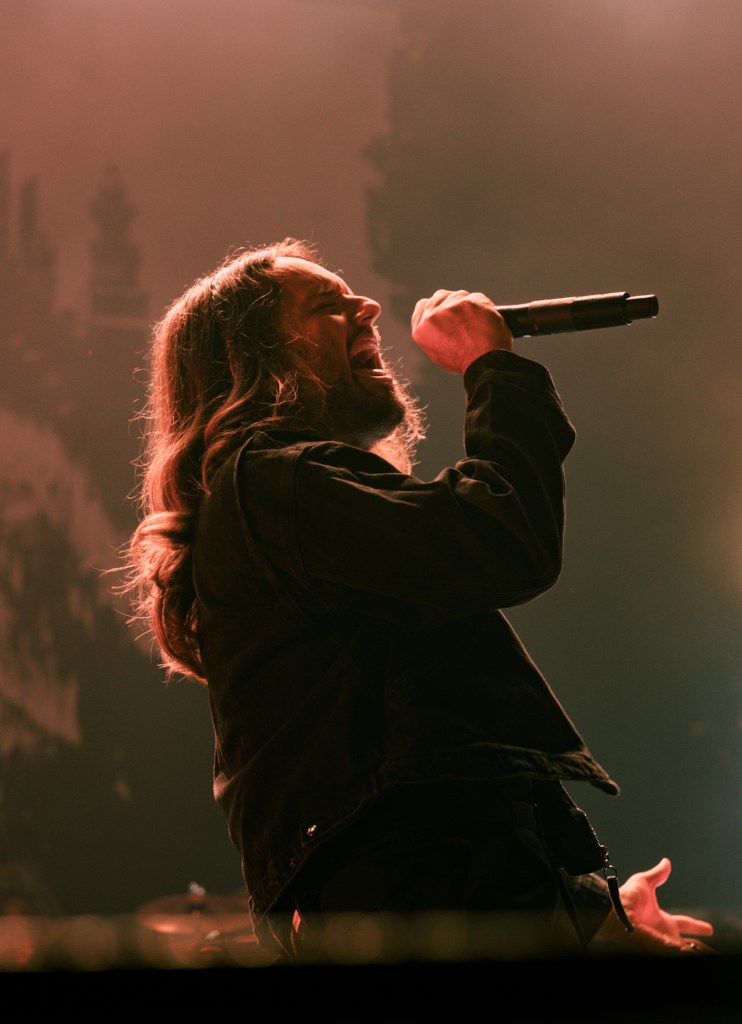A male vocalist with long hair passionately singing into a microphone on stage, surrounded by dramatic lighting.