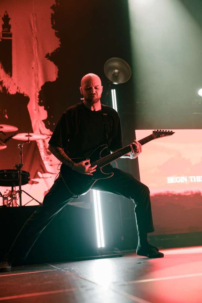 A guitarist performing energetically on stage with a dark background and a spotlight highlighting them. The scene captures the intensity of a live music event, with dramatic lighting and visual elements in the backdrop.