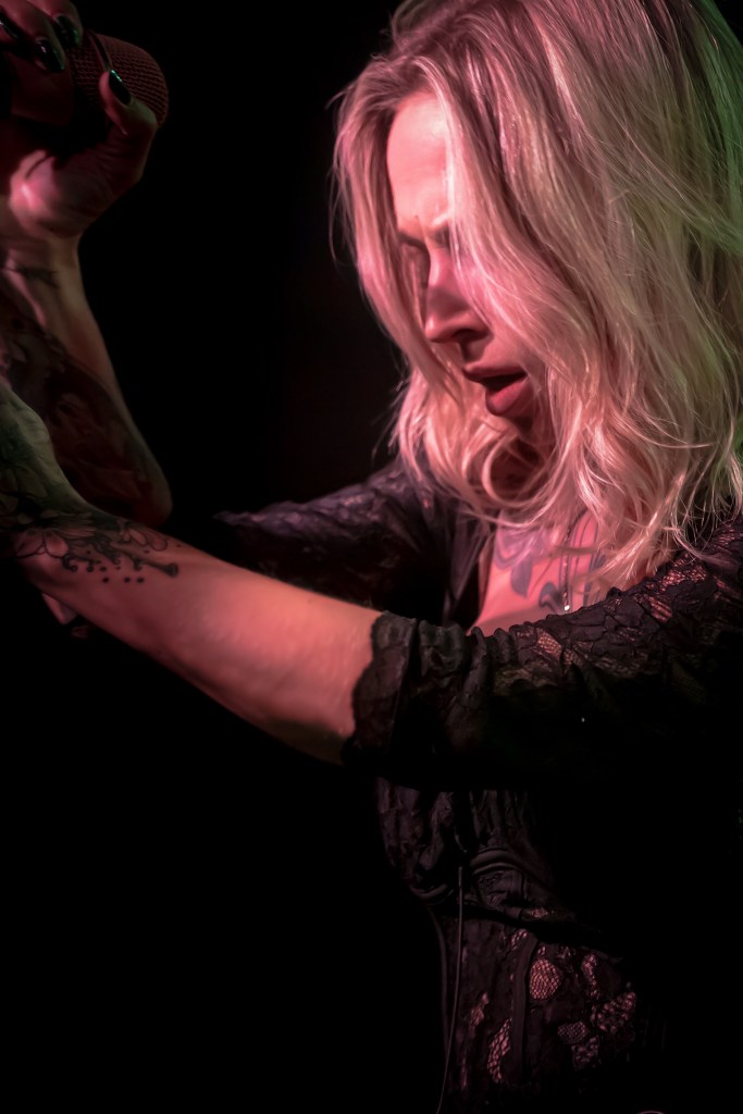 A close-up of a female singer with long blonde hair performing on stage, holding a microphone and passionately engaging with the audience.