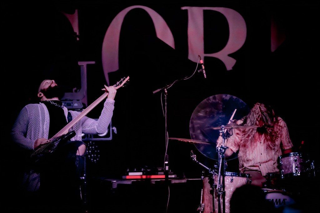 Two musicians performing on stage; one is playing an electric guitar while the other is drumming. The background features a partially visible band name.