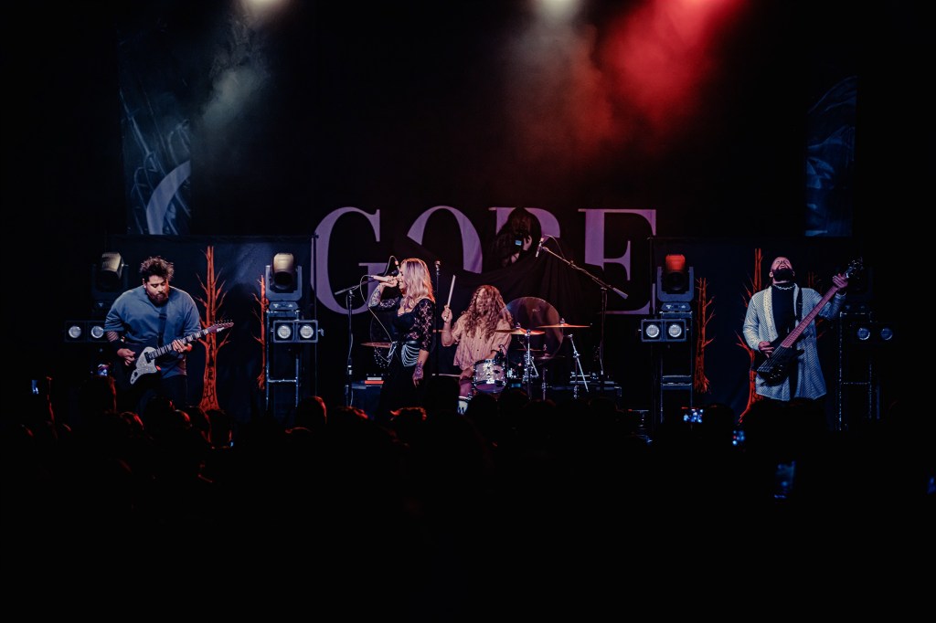A live music performance featuring a band on stage with a guitarist, vocalist, drummer, and bassist. The backdrop displays the word 'GORE' and colorful stage lights illuminate the scene.