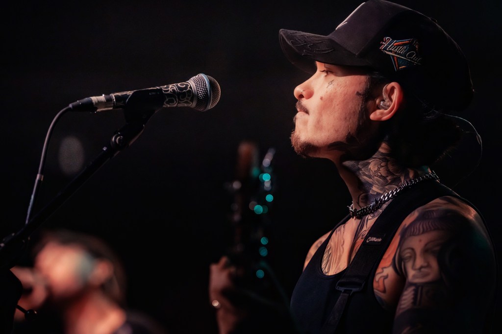 A side profile of a tattooed musician wearing a black cap, standing near a microphone while performing on stage.