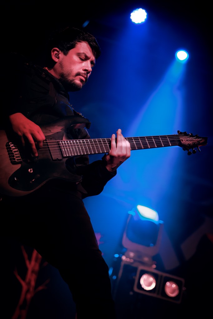 A musician playing an electric guitar on stage, illuminated by blue lights.