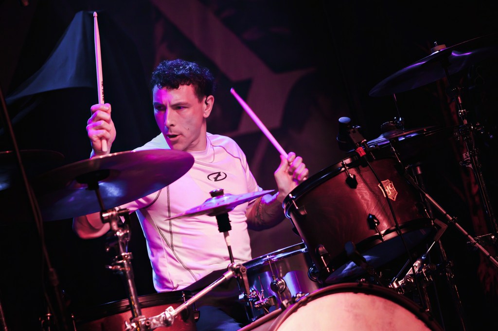 A drummer performing energetically on stage, playing a drum kit with drumsticks, wearing a fitted white shirt.