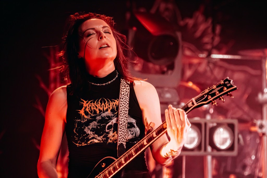 A female guitarist performing passionately on stage, wearing a black tank top with a graphic design, surrounded by dramatic lighting and stage equipment.