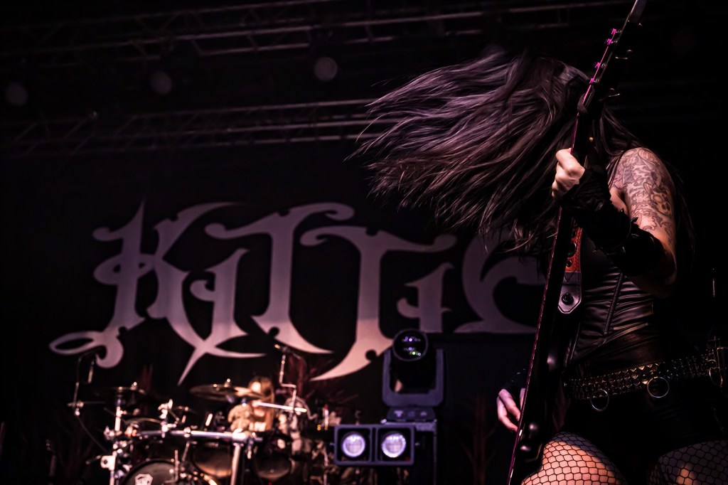 Female guitarist performing on stage with long hair swinging, wearing a dark outfit, in front of a large backdrop with the band name 'Kittie.'