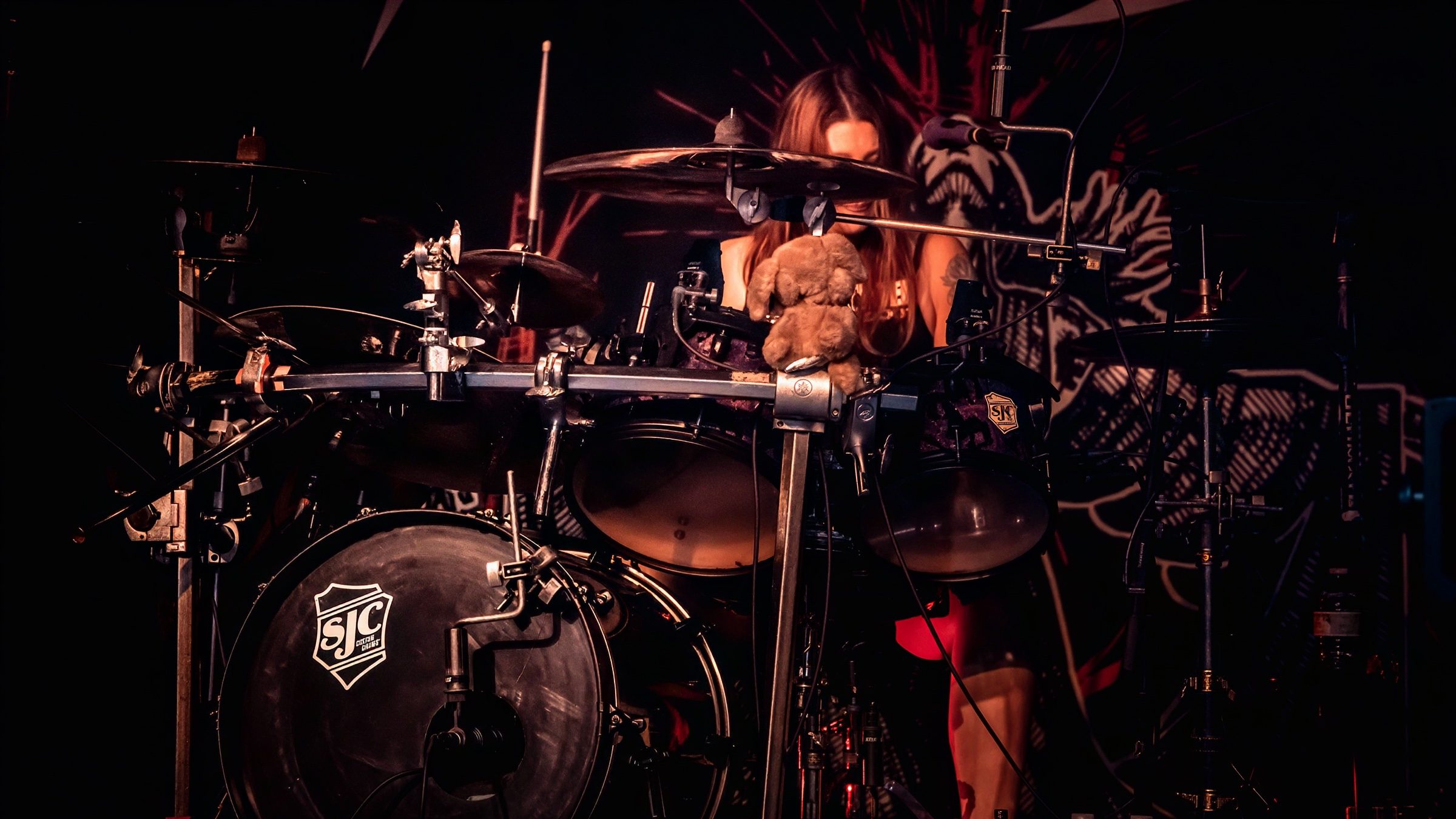 A drummer performing on stage with a stuffed animal on the drum kit, set against a colorful background.
