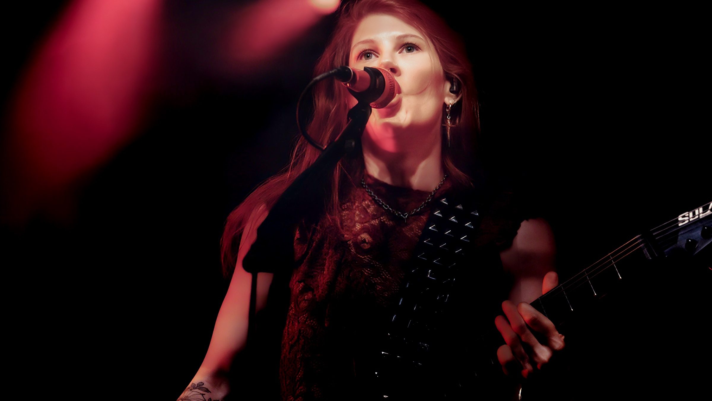 A singer performing on stage with a microphone, wearing a black outfit with spikes, illuminated by red stage lights.