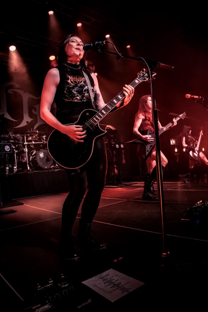 Female guitarist performing on stage with a microphone, wearing black clothing and playing an electric guitar, with band members in the background.