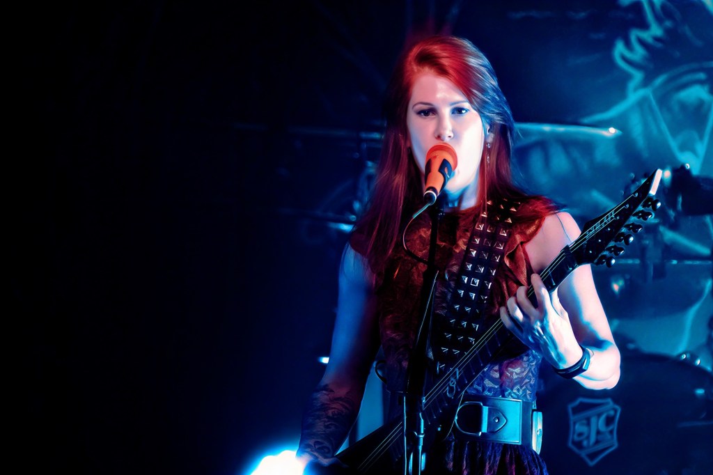 A female musician with red hair performing on stage, holding an electric guitar and singing into a microphone, with blue stage lighting in the background.