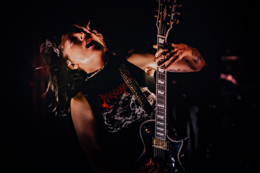 A guitarist passionately performing on stage, head thrown back and hair flying, wearing a black sleeveless shirt with a graphic design.