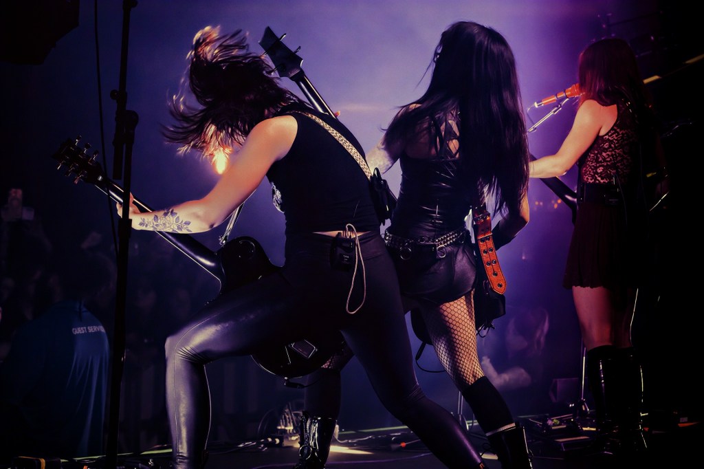 Three female musicians performing on stage with electric guitars, wearing stylish outfits and energetic poses under dramatic lighting.