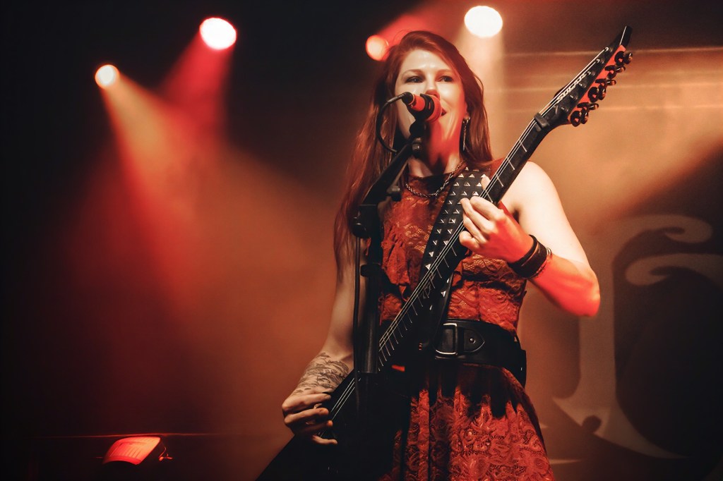 A female musician performing on stage with a microphone and electric guitar, wearing a red dress and adorned with accessories, surrounded by colorful stage lights.