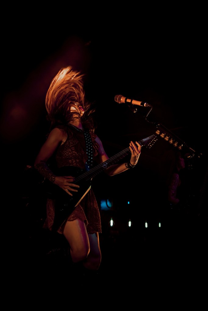 A female guitarist performing energetically on stage with her hair flying and a microphone in front of her, set against a dark background.