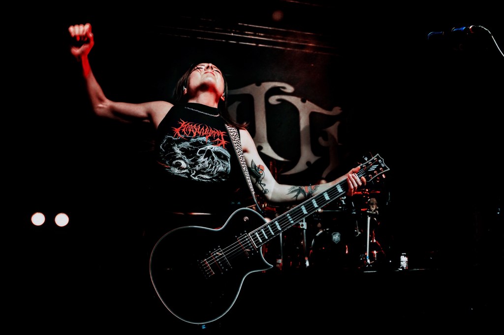 A musician playing electric guitar on stage, passionately performing with raised fists, wearing a black tank top featuring a graphic design.