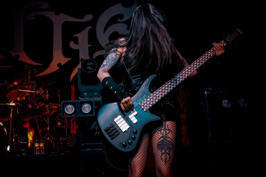 A female bassist performing on stage, wearing a black outfit and fishnet stockings, playing a blue bass guitar with long hair flowing as she moves.
