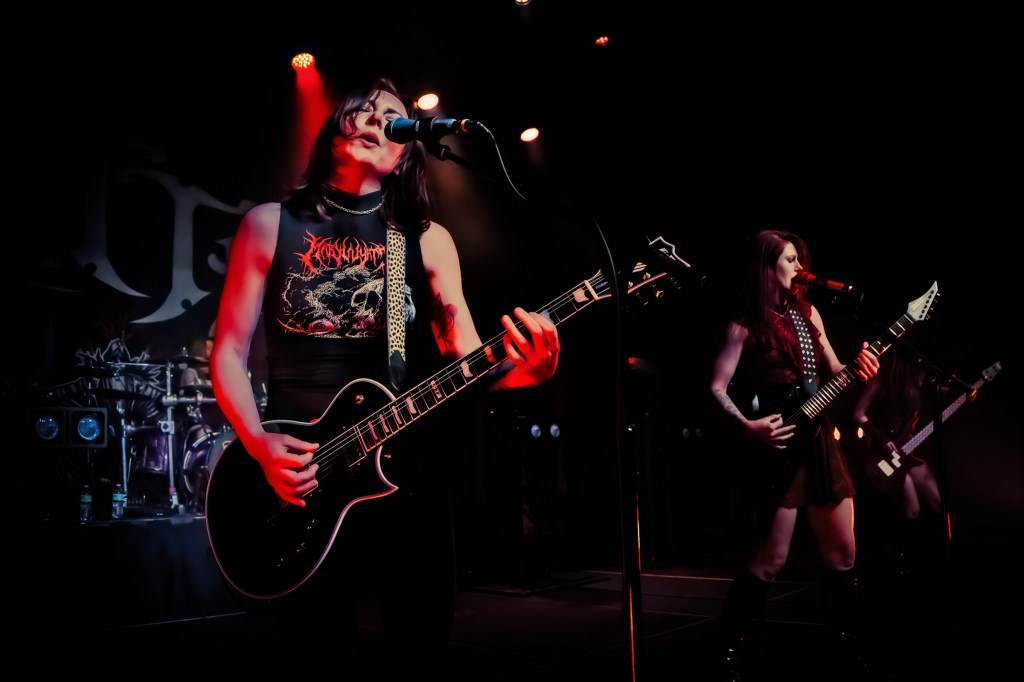 Two female musicians performing on stage with electric guitars and microphones, illuminated by colorful stage lights.
