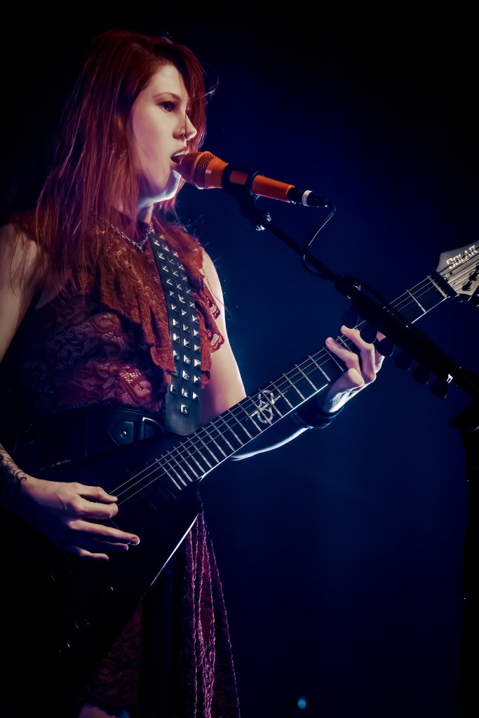 A female musician performing on stage, playing an electric guitar and singing into a microphone, dressed in a stylish outfit with a lace texture.