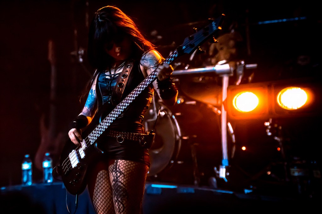 A female musician playing an electric bass guitar on stage, wearing a black outfit and fishnet stockings, with dramatic lighting in the background.