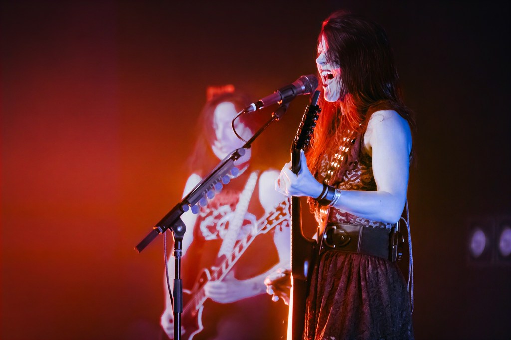 A female musician passionately singing into a microphone while playing an electric guitar on stage, with vibrant red and orange lighting in the background.