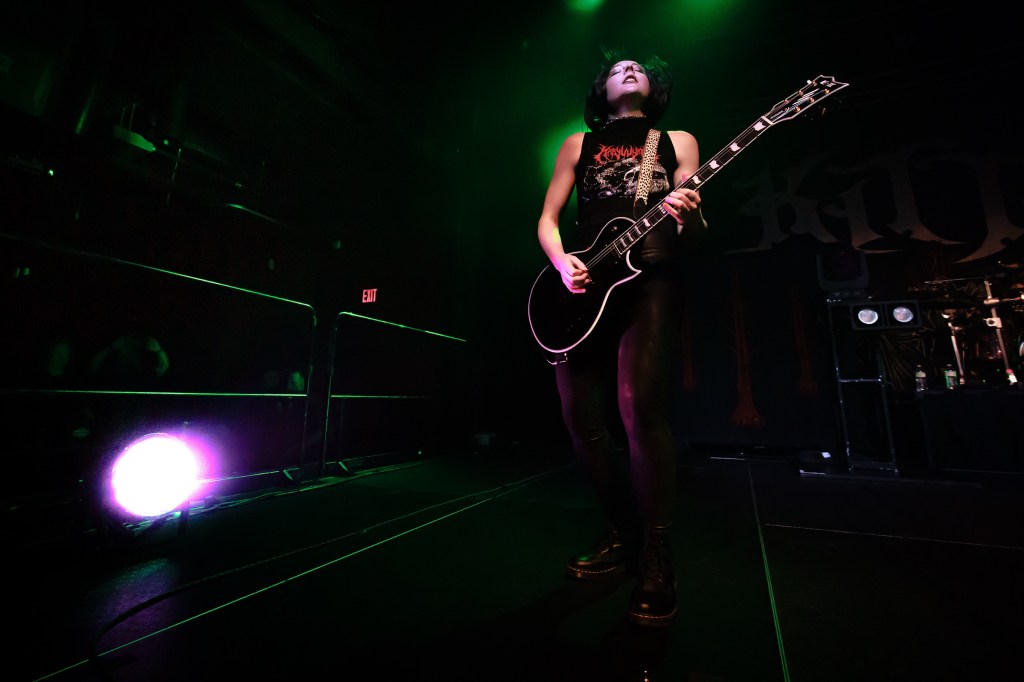 A guitarist performing on stage under green lighting, wearing a sleeveless shirt and black pants, with a focused expression and a black guitar.