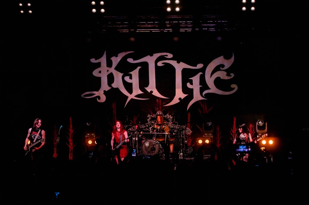 A live performance by the band Kittie on stage, featuring four band members playing instruments in front of a large Kittie logo backdrop.