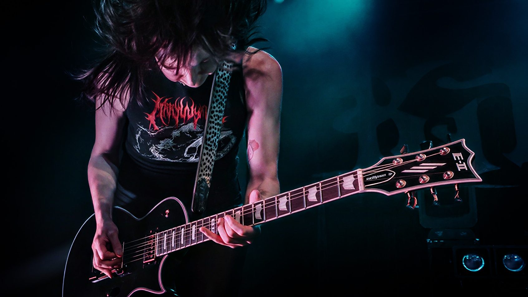 A guitarist passionately playing an electric guitar on stage, with long hair flowing and dramatic stage lighting in the background.